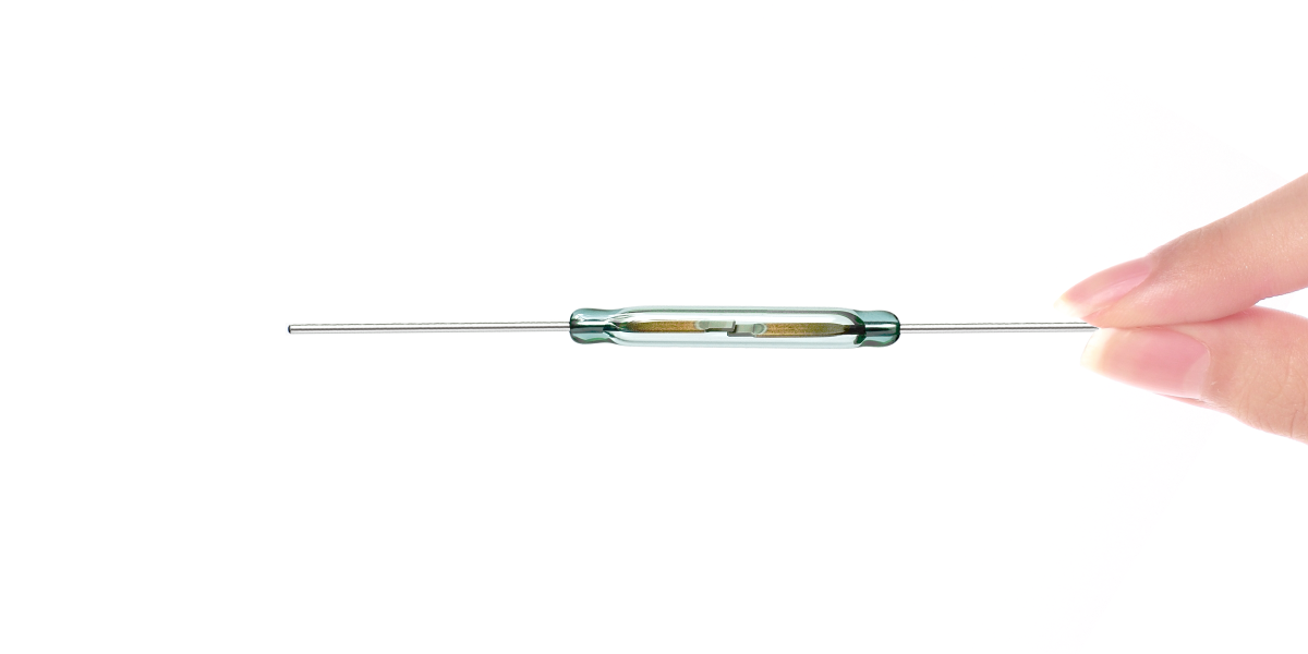 A hand holds a small, energy efficient sensor—a glass reed switch with metal leads extending from both ends—against a white background. by Standex Detect