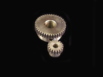 Two metal gears, one large and one small, positioned together on a black background, highlight the precision of engineering components often used alongside sensors to measure speed in mechanical systems. by Standex Detect